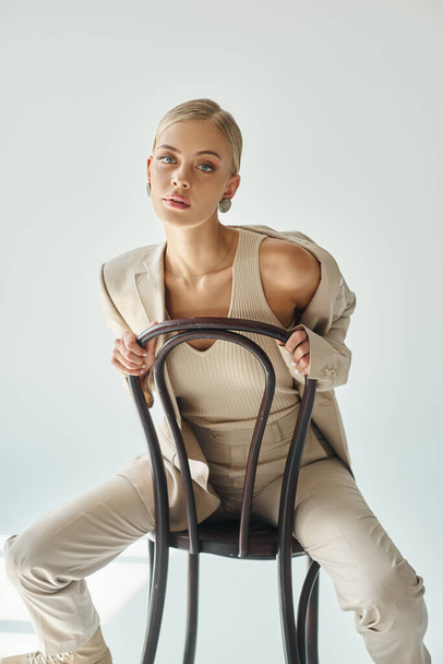 nuori Fashionista toppi ja beige puku katselee kameraa istuessaan tuoli harmaa - Valokuva, kuva