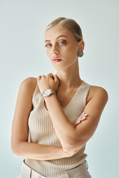 portrait of pretty blonde woman in beige tank top and wristwatch looking at camera on grey - Photo, Image