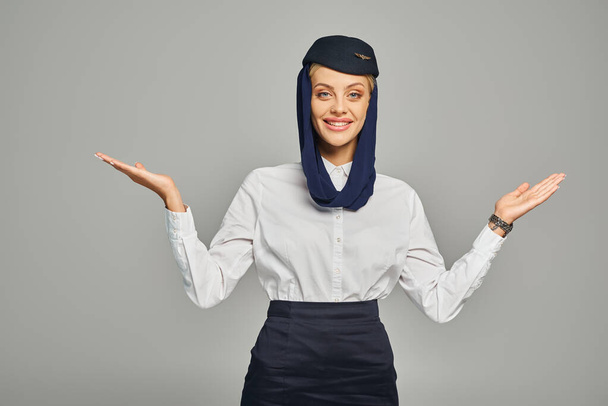 alegre azafata de las aerolíneas árabes en pañuelo para la cabeza y uniforme de pie con los brazos abiertos en gris - Foto, Imagen