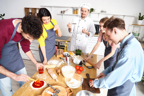 Ομάδα νέων με Ιταλό σεφ που ετοιμάζουν πίτσα κατά τη διάρκεια του μαθήματος μαγειρικής στην κουζίνα - Φωτογραφία, εικόνα
