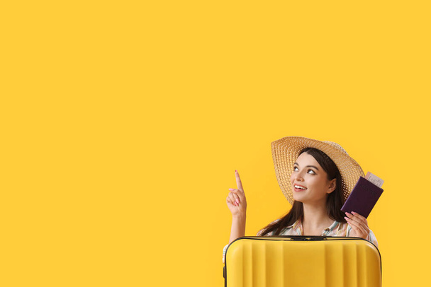 Young woman with passport and suitcase pointing at something on yellow background - Photo, Image