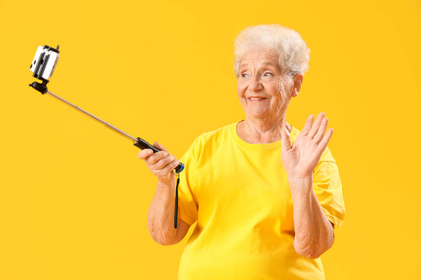 Starsza kobieta z telefonem komórkowym robi selfie na żółtym tle - Zdjęcie, obraz