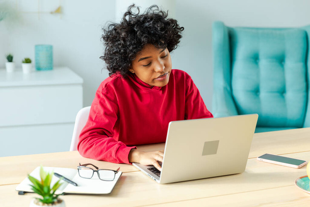 Egy afro-amerikai lány laptopot használ az otthoni irodájában, és a képernyőt nézi, amint chatel, miközben e-maileket ír. Fiatal nő, hogy virtuális találkozó online chat video call konferencia. Otthoni tanulás - Fotó, kép