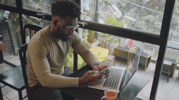 Tiro de ángulo alto del joven empresario afroamericano en ropa de casualwear y mensajes de gafas en el teléfono inteligente mientras está sentado con el ordenador portátil y taza de té en la mesa en la cafetería - Imágenes, Vídeo