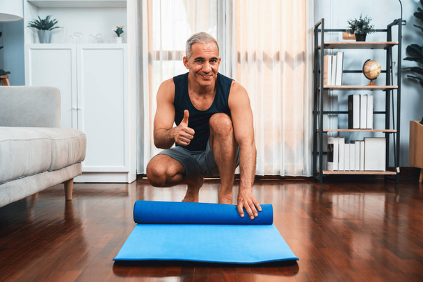Активний і спортивний старший чоловік готує, катається фітнес тренувальний килимок на підлозі у вітальні вдома. Домашні вправи як концепція здорового способу життя тіла після виходу на пенсію. Розрізати - Фото, зображення