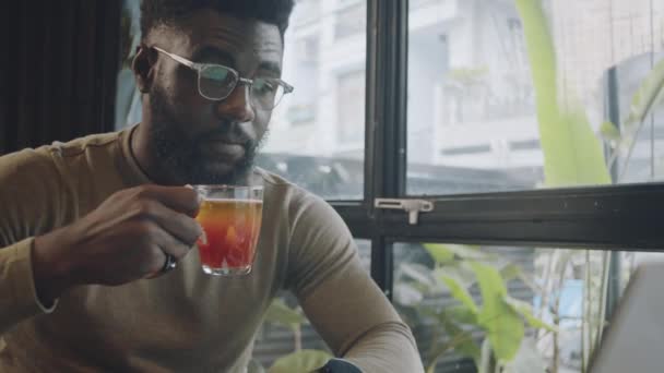 Genç Afro-Amerikan iş adamının günlük kıyafetleri ve gözlükleriyle çay içerken ve kafede uzaktan çalışırken dizüstü bilgisayar ekranına bakarken orta boy yakın çekimi. - Video, Çekim