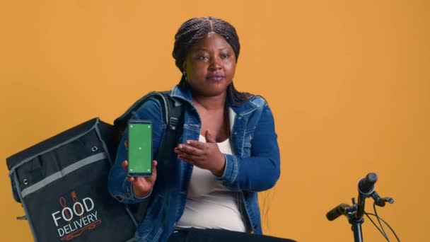 Γυναικεία αφρικάνικη αμερικανική courier κρατώντας ψηφιακή συσκευή με πράσινη οθόνη, ενώ κάθεται στο ποδήλατο. Νεαρή γυναίκα παράδοση δείχνει smartphone με κενό πρότυπο copyspace mockup. - Πλάνα, βίντεο
