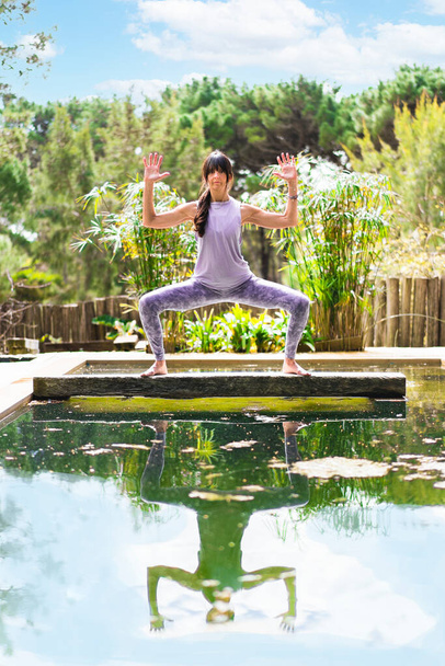 Una mujer practicando la pose de yoga diosa con brazos de cactus en un tronco de árbol sobre una piscina, tiro vertical - Foto, imagen