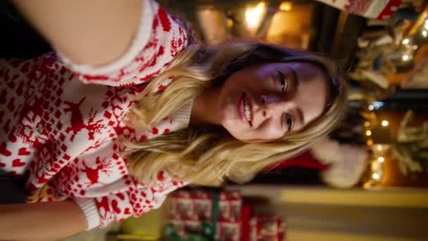 Πανέμορφη ξανθιά γυναίκα που φοράει χειμερινό πουλόβερ απολαμβάνοντας βιντεοκλήση στο smartphone με χαμόγελο ευτυχία. Όμορφο κορίτσι κουβεντιάζοντας με web κάμερα στο χριστουγεννιάτικο δέντρο και φώτα bokeh pov κάθετο υλικό 4K - Πλάνα, βίντεο