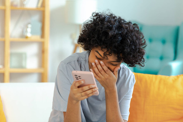 Innoissaan onnellinen nuori musta afrikkalainen amerikkalainen nainen tilalla matkapuhelin nauraa tunne iloa saada mobiili viesti. Iloinen tyttö nauraa ääneen istuu sohvalla katsomassa hauska video käsittelyssä uutisia - Valokuva, kuva
