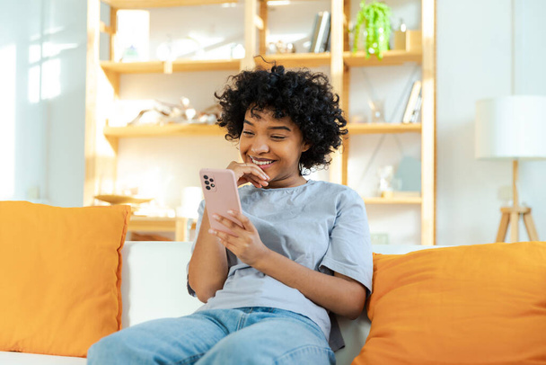 Вражена щаслива молода чорношкіра американка, яка тримає мобільний телефон, сміючись, отримує задоволення від мобільного повідомлення. Захоплива дівчина сміється вголос сидячи на дивані і дивиться смішні новини з читання відео - Фото, зображення