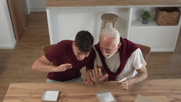 senior man grandfather pensioner gray hair and beard with his grandson teenager boy  receive presents in box open read card happy smile in front of digital tablet at home online video call slow motion - Footage, Video
