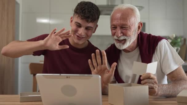 senior man grandfather pensioner gray hair and beard with his grandson teenager boy  receive presents in box open read card happy smile in front of digital tablet at home online video call slow motion - Footage, Video
