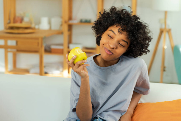 Happy pretty girl biting green apple at home. Beautiful african american young woman eating fresh fruit and smiling. Healthy food vegan vegetarian dieting concept. Healthy snack clean food - Photo, Image