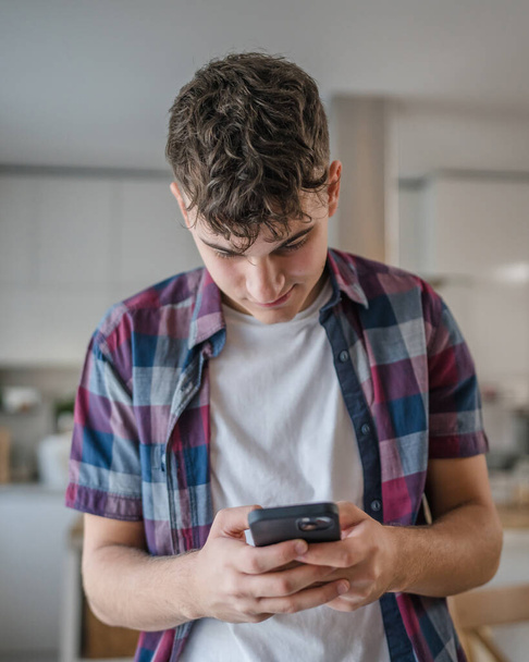 Ένας άνδρας καυκάσιος έφηβος αγόρι χρησιμοποιούν κινητό τηλέφωνο smartphone για online πλοήγηση στο διαδίκτυο κοινωνικό δίκτυο ή sms μηνύματα κειμένου στο σπίτι χαρούμενος χαμόγελο - Φωτογραφία, εικόνα
