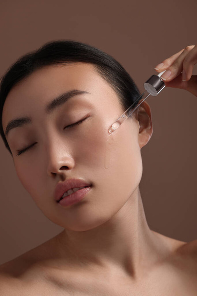 Beautiful young woman applying cosmetic serum onto her face on brown background, closeup - Valokuva, kuva