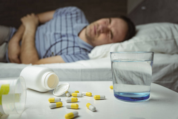 A man in home clothes and holds his hands on his stomach lying on the bed. the table, on which there are tablets, pills and a glass of water. Severe abdominal pain, malaise, cold. - Photo, Image