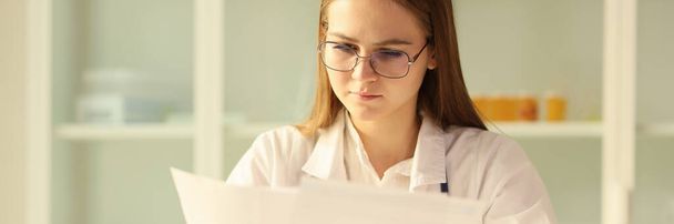 Gözlüklü genç doktor klinikteki masada otururken tıbbi belgeleri okuyor. Hemşire hastanede evrak işi yapıyor.. - Fotoğraf, Görsel