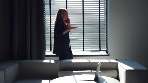 Wütende Frau im schicken Anzug schreit und gestikuliert während eines Handy-Gesprächs mit einem Geschäftspartner, während sie am Fenster steht. Sonnenlicht im dunklen Büro durch geöffnete Jalousien. - Filmmaterial, Video
