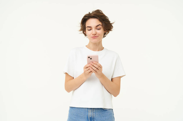 Πορτρέτο της μελαχρινής κοπέλας με smartphone, αποστολή μηνύματος, χρήση εφαρμογής κινητού τηλεφώνου, χαμόγελο και εμφάνιση χαρούμενη, λευκό φόντο στούντιο. - Φωτογραφία, εικόνα
