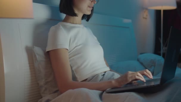 Vágott felvétel nő ül az ágyon este otthon, és gépelés a laptopon - Felvétel, videó
