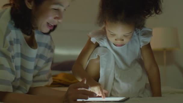 Χαμογελώντας ασιατική μητέρα και αξιολάτρευτο κοριτσάκι παίζουν μαζί σε ψηφιακή ταμπλέτα το βράδυ στο σπίτι - Πλάνα, βίντεο