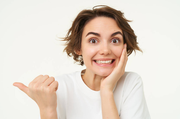 Immagine di donna bruna eccitata con taglio di capelli corto, indicando a sinistra, sorridente e guardando felice, mostrando pubblicità, in piedi su sfondo bianco. - Foto, immagini