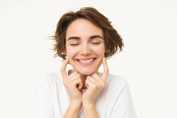 Portrait of brunette woman smiling, shuts her eyes and touches dimples on her cheeks, stands over white background. Copy space - Fotografie, Obrázek