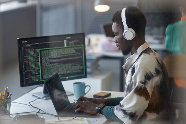 Αφροαμερικανός προγραμματιστής σε ακουστικά πληκτρολογώντας στο laptop στο χώρο εργασίας του - Φωτογραφία, εικόνα