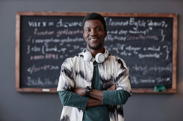 Porträt eines afroamerikanischen Lehrers, der in die Kamera lächelt, während er mit verschränkten Armen und einer Tafel im Hintergrund steht - Foto, Bild