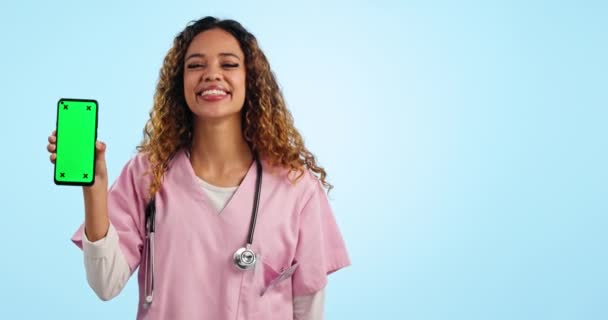 Portrét ženského lékaře ukazujícího na telefon se zelenou obrazovkou, maketou a sledovacím značkovačem ve studiu. Úsměv, telehealth a šťastný zdravotní sestra s smartphone chromakey, zpětná vazba nebo webové stránky na modrém pozadí. - Záběry, video