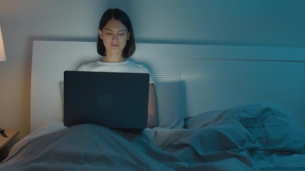 Medium shot van jonge vrouw zitten op bed thuis en werken op laptop voor het gaan slapen 's nachts - Video