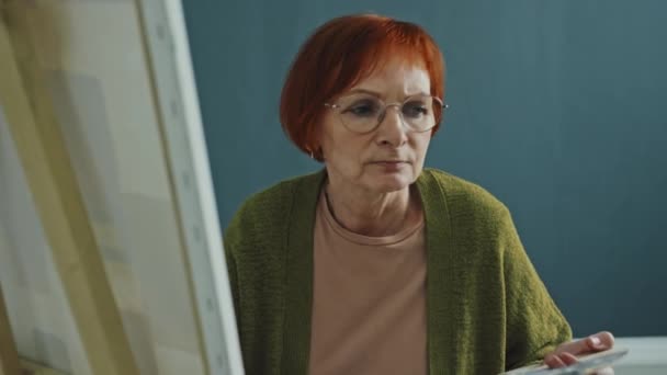 Średnie ujęcie starszej kobiety w czerwonych włosach noszącej okulary, trzymającej paletę i pędzel w dłoniach siedzącej przed płótnem i rysującej - Materiał filmowy, wideo