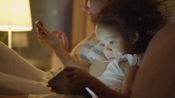 Mittlere Nahaufnahme eines entzückenden asiatischen Kleinkindes, das Cartoon auf einem digitalen Tablet anschaut, während die Mutter abends zu Hause im Bett telefoniert - Filmmaterial, Video