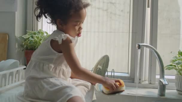 Портативная камера, на которой мама-азиатка полоскает тарелку чистой водой, как милая маленькая дочь сидит у раковины, играет с ковшами и помогает мыть посуду на кухне дома - Кадры, видео