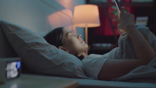 Mujer joven usando el teléfono móvil en la cama, luego acostado debajo de la manta y quedarse dormido por la noche - Metraje, vídeo
