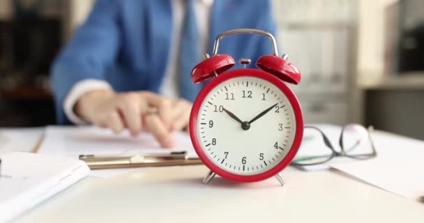 Reloj despertador clásico rojo de pie delante de un hombre de negocios estudiando documentos 4k película. Concepto de gestión del tiempo - Imágenes, Vídeo