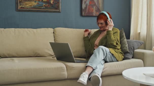 Panning right full shot of cheerful senior woman sitting on sofa with laptop saying hi to relatives during video call - Footage, Video