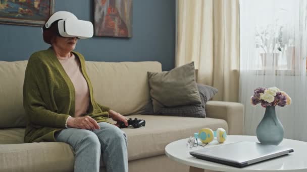 Medium full shot of senior Caucasian woman sitting on couch in living room feeling tired after playing games in AR goggles taking them off - Footage, Video