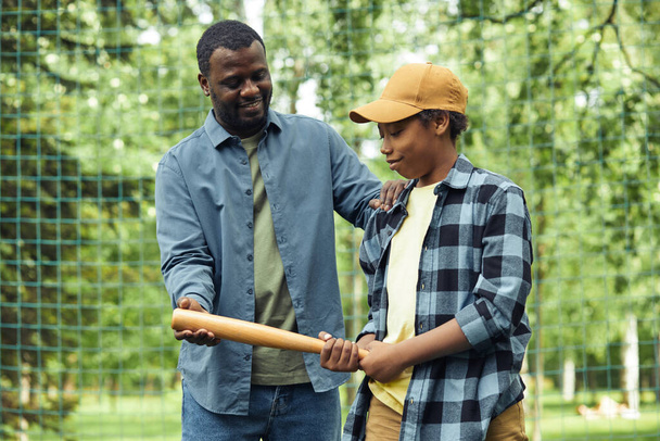 Afrikanischer junger Vater gibt seinem Sohn den Schläger und zeigt ihm, wie man ihn hält, während er ihm Baseball beibringt - Foto, Bild