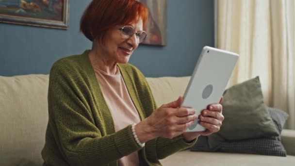 Medium shot of smiling senior Caucasian woman sitting on sofa in living room holding tablet waving hand and talking to family via video call - Video, Çekim