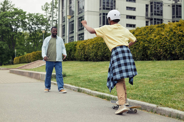 Afrikanischer Junge mit Helm fährt mit seinem Vater auf Skateboard auf der Straße, während er ihn unterrichtet - Foto, Bild
