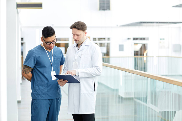 Portrait of two doctors looking at clipboard while standing at balcony in modern clinic interior, copy space - Photo, Image