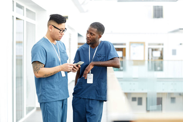 Portrait de deux médecins multiethniques portant l'uniforme debout dans le hall de la clinique moderne et bavardant, espace de copie - Photo, image