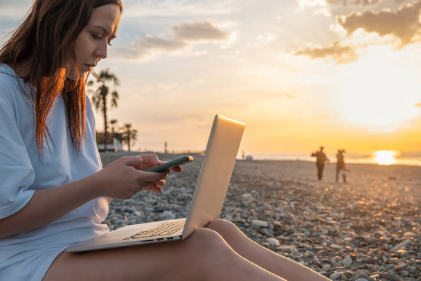 Günbatımında sahilde dizüstü bilgisayar ve cep telefonuyla çalışan dijital göçebe kadın. Serbest çalışan bir kadın uzaktan akıllı telefon ve bilgisayar kullanarak çevrimiçi çalışıyor. Tatilde uzaktan çalışma - Fotoğraf, Görsel