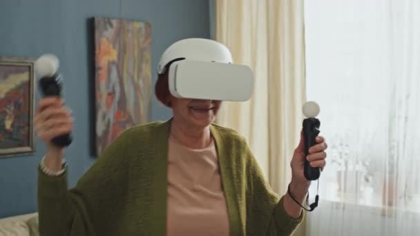 Primer plano medio de la mujer mayor con gafas AR y sensores de movimiento de retención jugando juegos en medio del apartamento - Metraje, vídeo