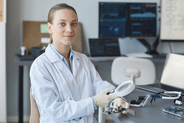 Πορτρέτο της νεαρής γυναίκας επιστήμονα χαμογελώντας στην κάμερα, ενώ κάθεται στο χώρο εργασίας στο εργαστήριο μηχανικής, αντίγραφο χώρο - Φωτογραφία, εικόνα
