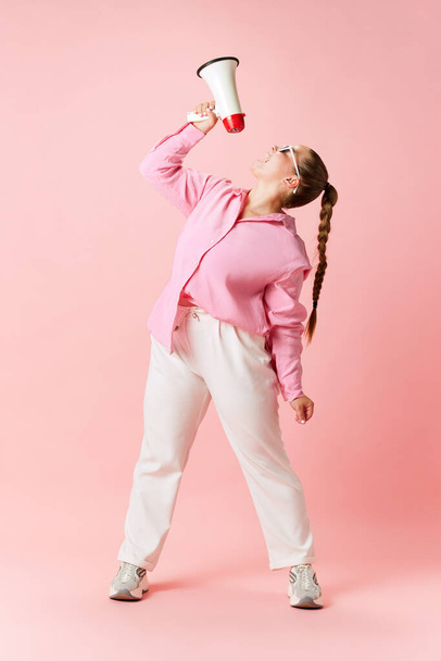 Full length portrait of plus size woman posing in stylishly casual outfit, in glasses shouting to megaphone of beginning sales season against pink background. Concept of fashion, business, shopping. - Foto, imagen
