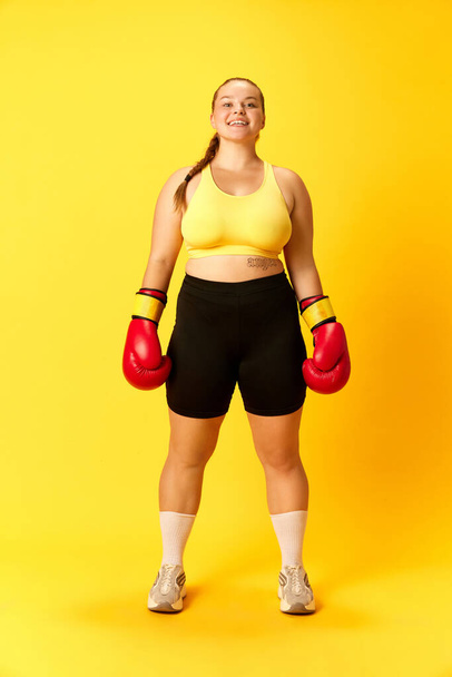 Portrait pleine longueur d'une jolie femme souriante de taille plus posant en tenue de sport, avec des gants de boxe sur fond jaune. Concept de sport, mode, passe-temps, temps libre, mode de vie sain. Publicité - Photo, image