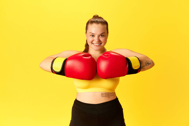 Image d'une jeune sportive joufflue en survêtement et rouge en gants de boxe mettant les mains ensemble isolées sur fond jaune. Concept de sport, passe-temps, mode de vie sain, entraînement, entraînement. publicité - Photo, image